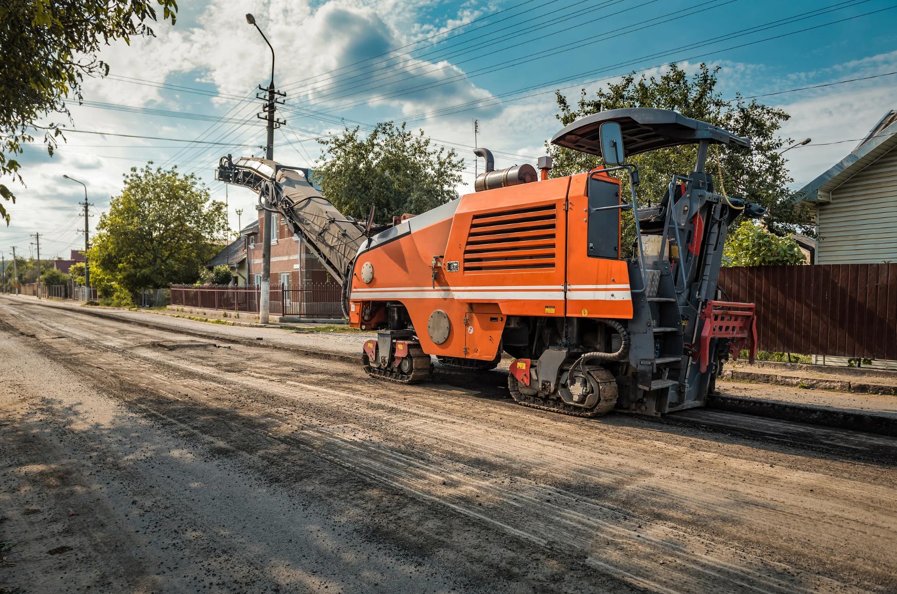 asphalt-milling-img-3 Asphalt Milling: An orange road milling machine removes old asphalt from a residential street, surrounded by houses, trees, and utility poles under a partly cloudy sky.