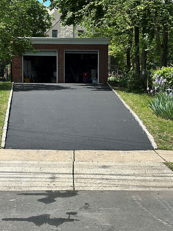 Freshly paved driveway leading up to a two-car brick garage with trees and plants on either side.