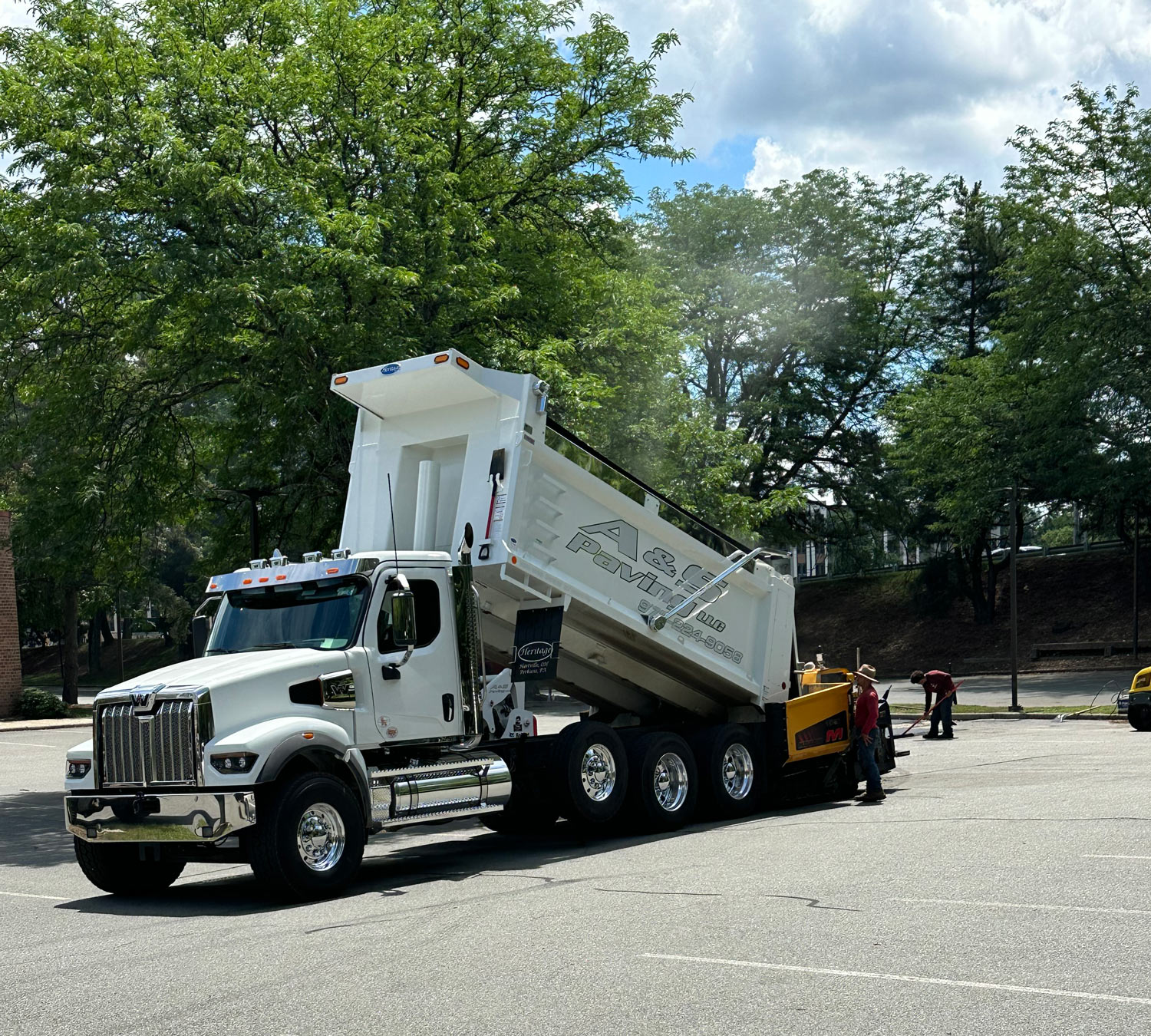 image0 Paving Services: Workers pave a road; one spreads asphalt while another drives a roller. A yellow paving machine lays fresh asphalt, and trees with autumn leaves are visible in the background.