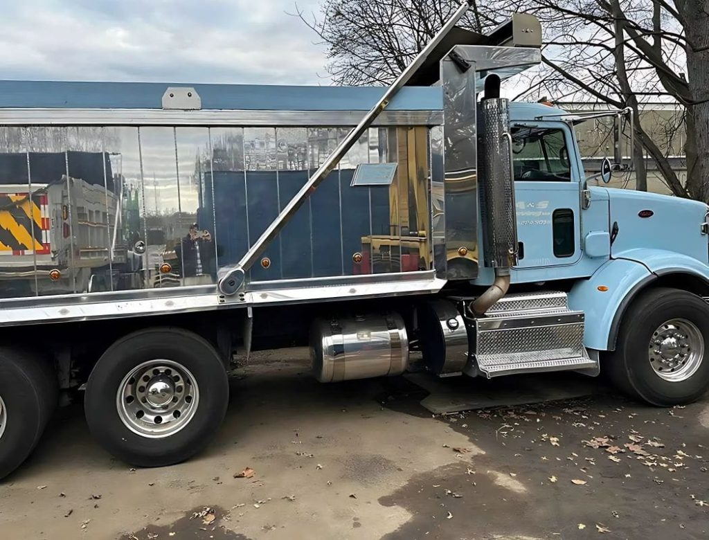 A shiny light blue dump truck is parked on a street beside leafless trees and scattered leaves.
