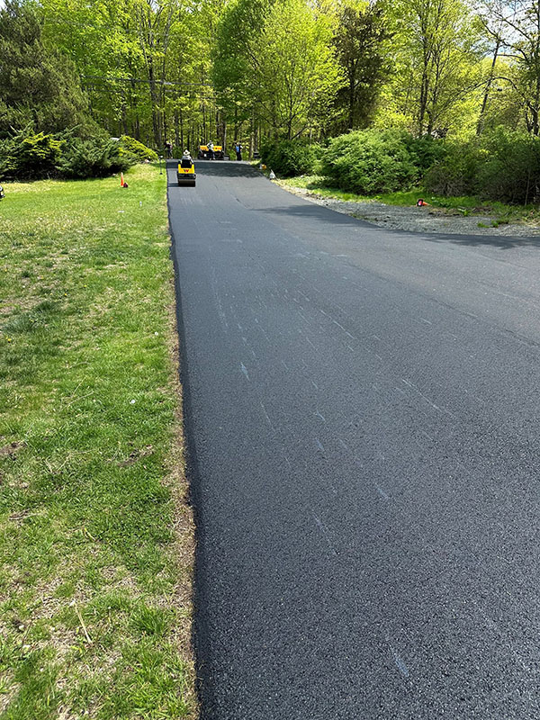 Freshly paved asphalt road through green trees with a yellow roller machine in the distance.