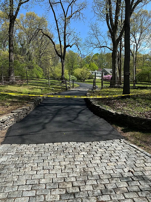 A paved driveway blocked by yellow caution tape, with trees and a house visible in the background.
