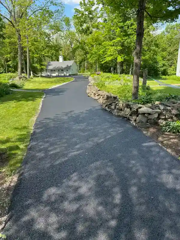 Freshly paved asphalt driveway curves through green yard toward a house, bordered by a stone wall and trees.