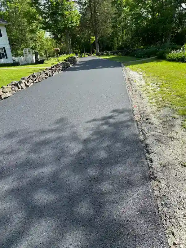 Freshly paved asphalt driveway bordered by a stone wall on one side and grass on the other, on a sunny day.