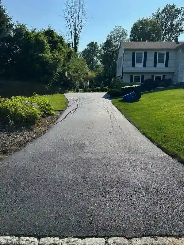 A newly paved driveway curves toward a white house with green lawn and trees in the background.
