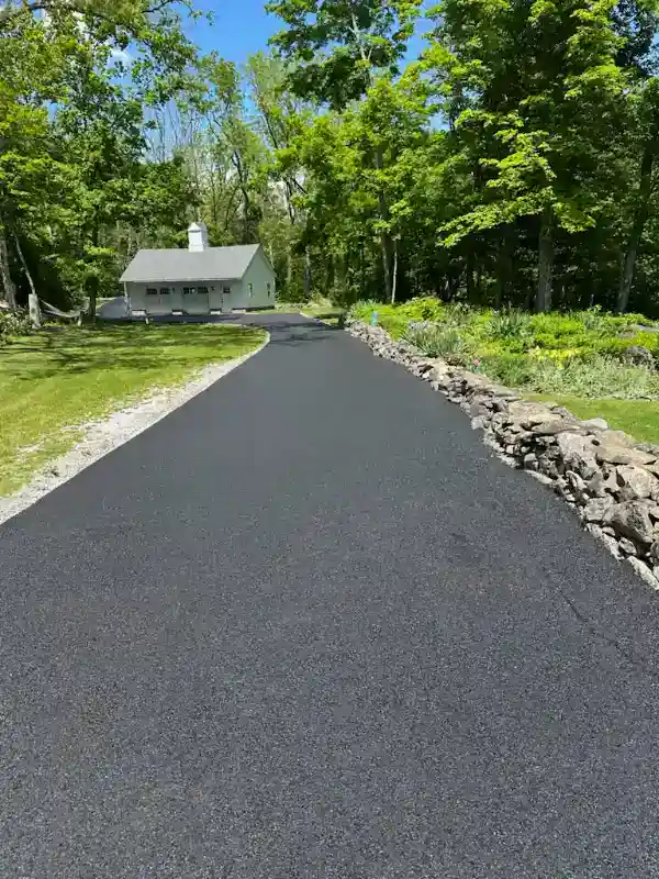 Freshly paved black asphalt driveway leading to a white house surrounded by green trees and grass.