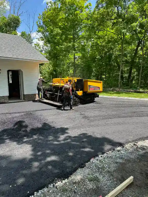 Two workers use a yellow asphalt paving machine to lay new pavement outside a white house on a sunny day.