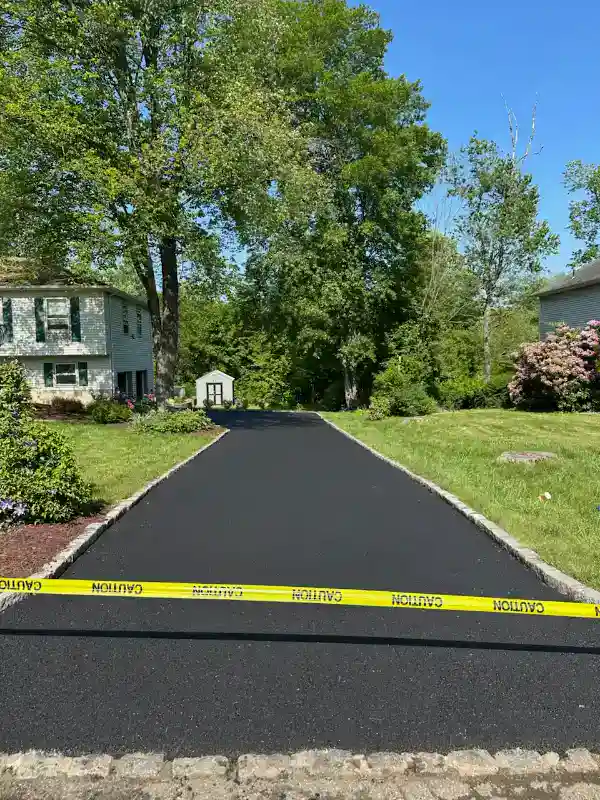 Freshly paved driveway with yellow caution tape across and houses on either side, surrounded by green trees.