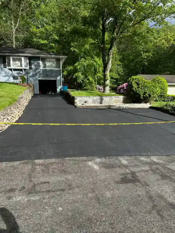 Freshly paved driveway in front of a house, blocked off with yellow caution tape, surrounded by greenery.