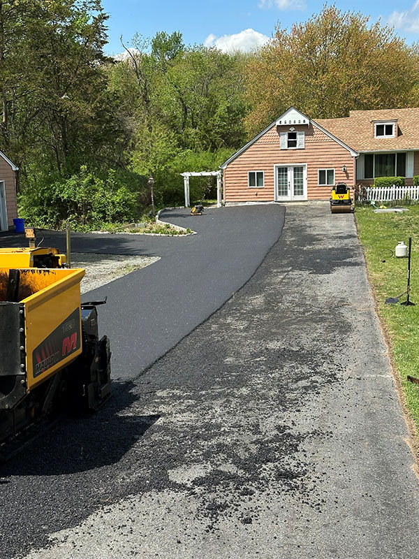 Fresh asphalt being laid on a driveway in front of a house, with paving machinery visible.