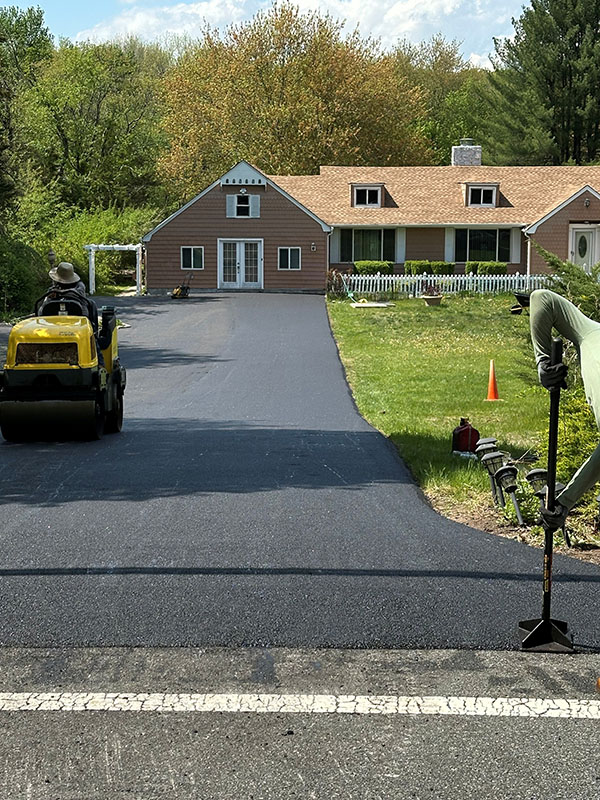 Workers pave a driveway in front of a house with a lawn and trees in the background.