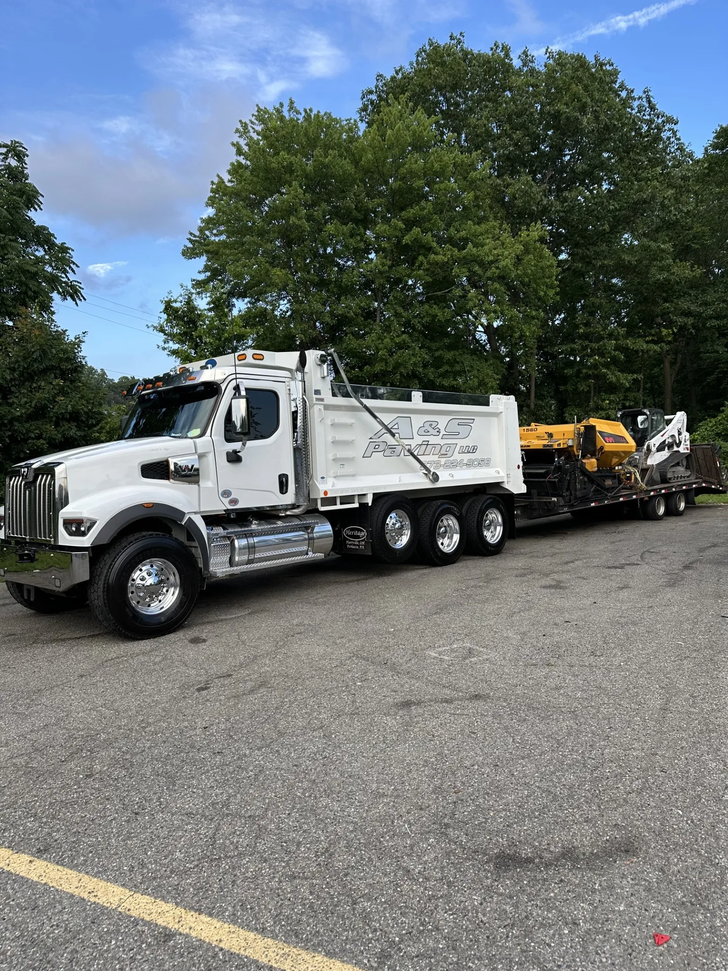 AS Truck 1 A newly paved asphalt driveway curves through a green lawn toward a white house with black shutters, surrounded by trees and a rock-lined garden under a blue sky with scattered clouds.