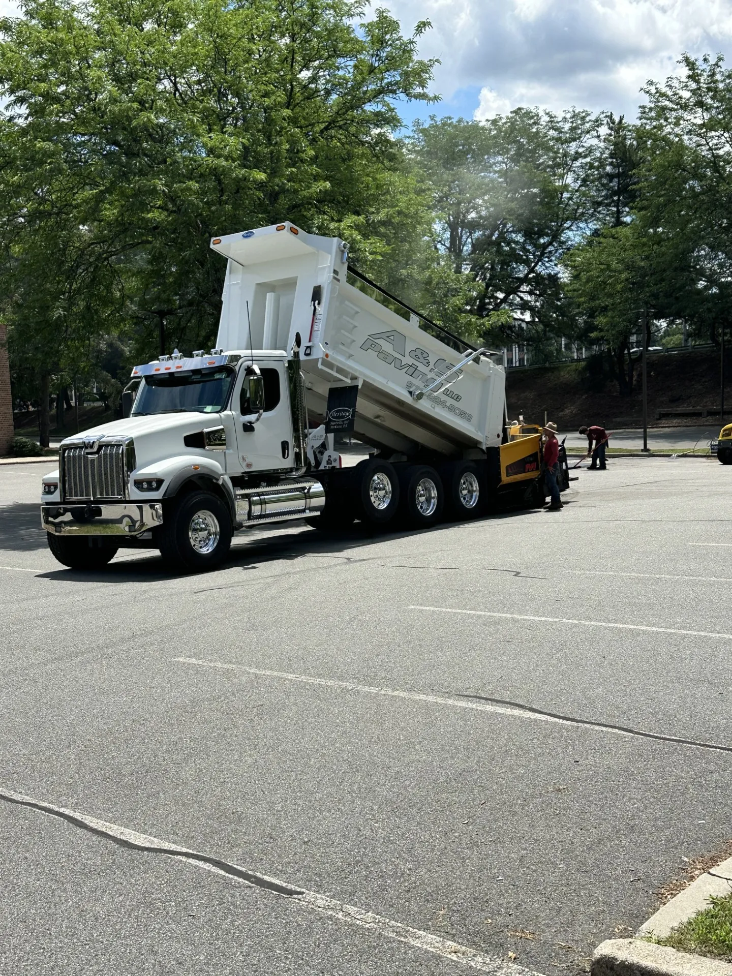 AS Truck 2 A&S Paving: A newly paved asphalt driveway curves through a green lawn toward a white house with black shutters, surrounded by trees and a rock-lined garden under a blue sky with scattered clouds.