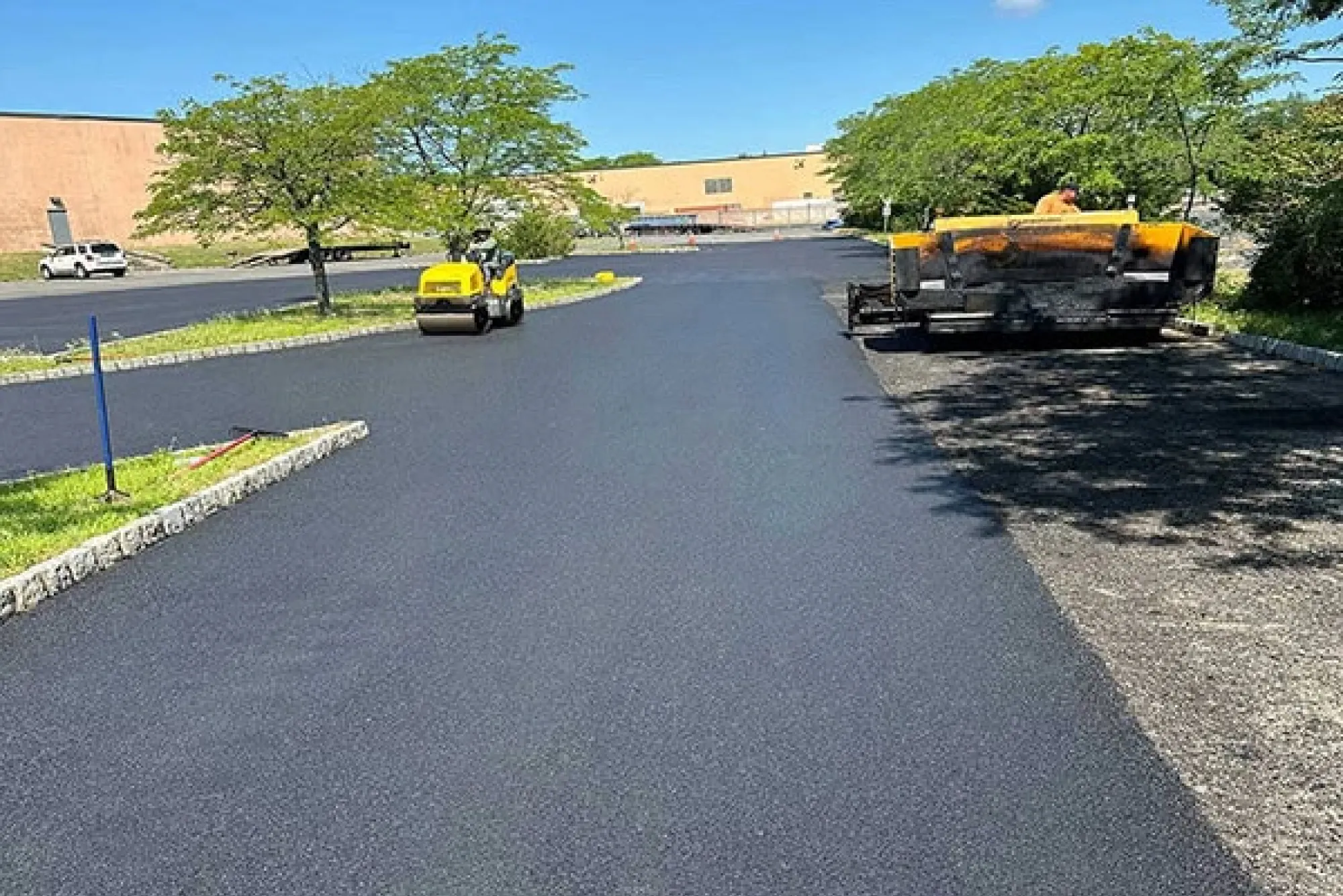 Fresh asphalt being laid in a parking lot with paving machinery and a worker under a clear blue sky.