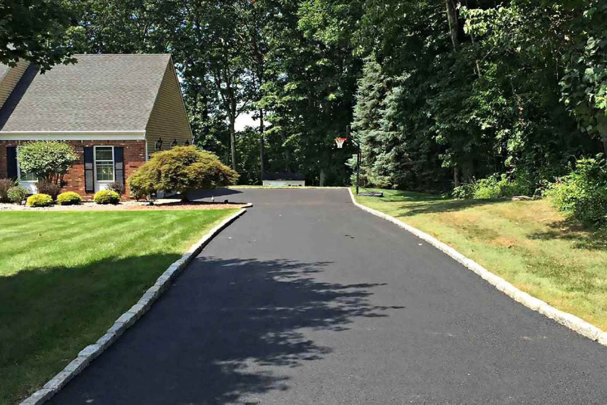 A newly paved black asphalt driveway curves past a brick house with green lawns and landscaping, bordered by stone edging and surrounded by tall trees.