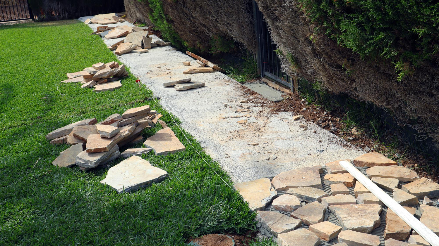 Broken stone slabs scattered along a sidewalk next to grass and hedges, with some stones partially arranged.