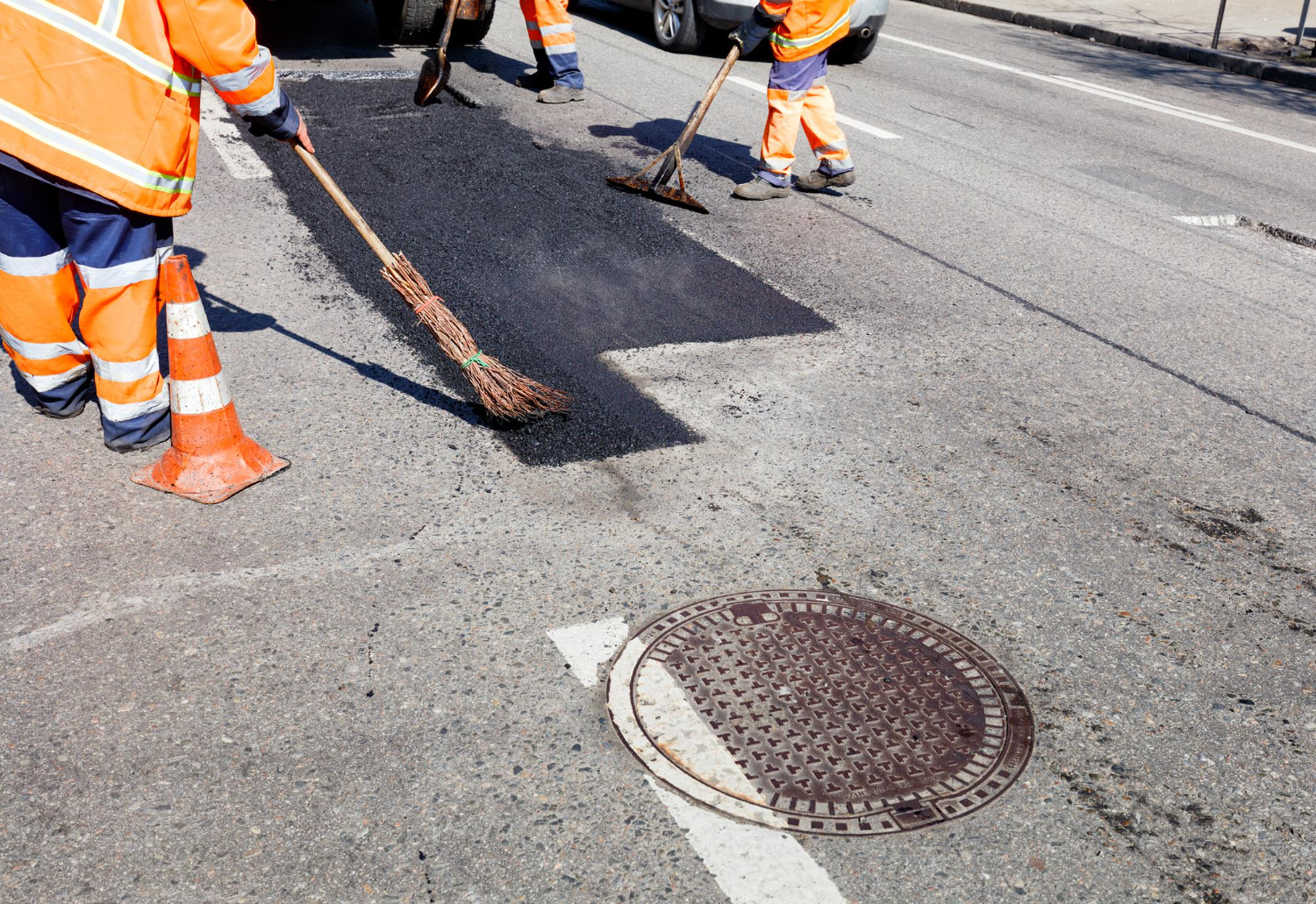 28596210 Workers in orange uniforms repair a road, sweeping new asphalt near a traffic cone and manhole cover.