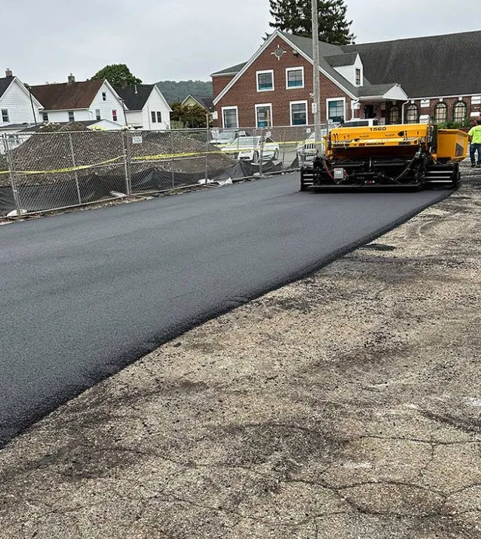 A yellow paving machine lays fresh asphalt on a road near houses and a fenced construction site.